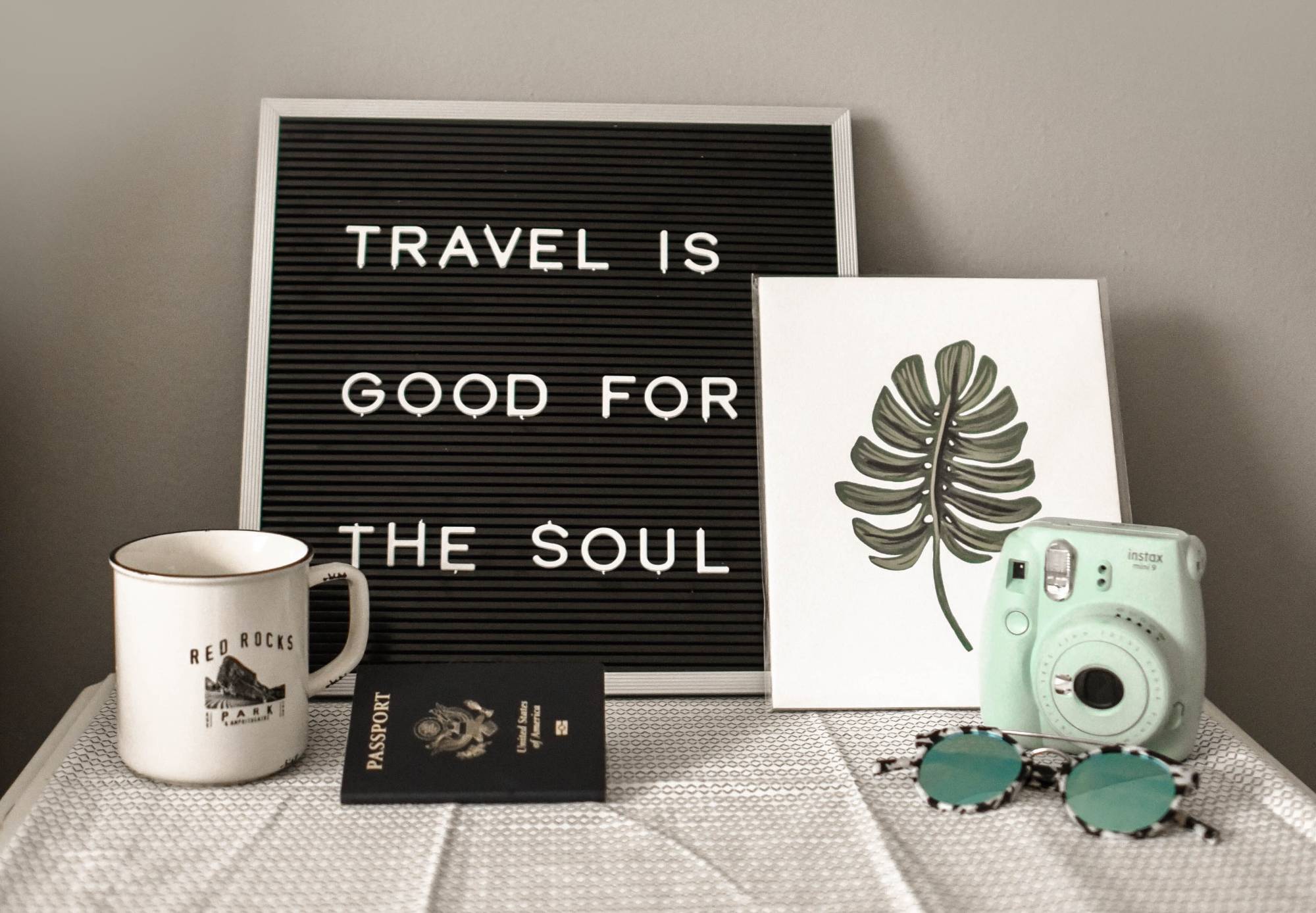 Sign stating Travel is good for the Soul along with a mug, passport, leaf print, camera, and sunglasses